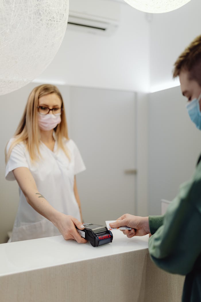 services-04 A masked patient pays at a clinic counter, using a card terminal handled by a healthcare professional.